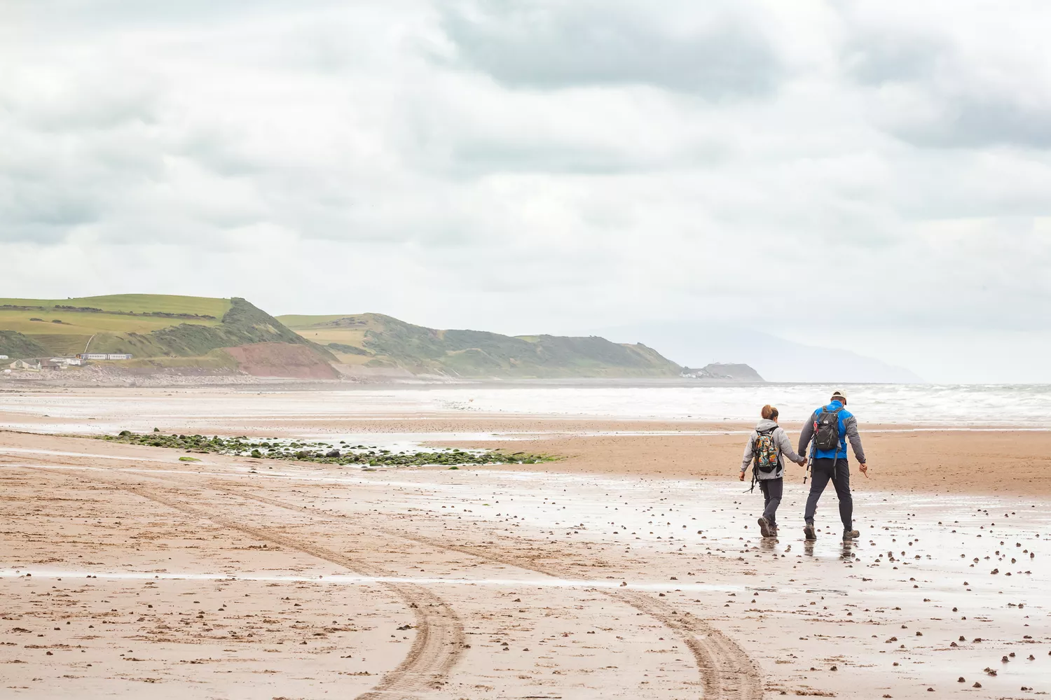 Two individuals wander the sandy beach, backpacks slung over their shoulders, embracing the coastal scenery.